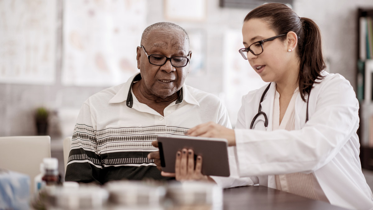 A doctor is sitting with a senior citizen to point out important information about health coverage on a digital tablet.