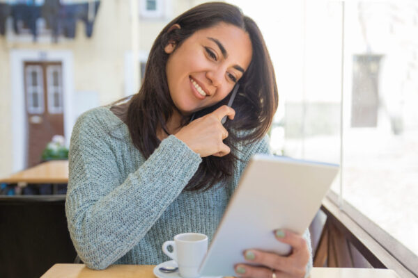 Smiling beautiful young woman using smartphone and tablet. Lady calling on mobile phone and browsing on tablet computer in cafe. Technology and communication concept. Front view.