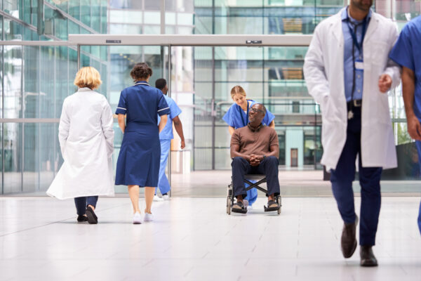 Female Nurse Wearing Scrubs Wheeling Patient In Wheelchair Through Lobby Of Modern Hospital Building